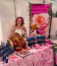 Load image into Gallery viewer, Enchanting Flowers founder Rosanne at a holistic wellbeing event, showcasing her handmade natural essential oil Aura Sprays, oracle cards and spiritual gifts The setup includes a large pink floral banner reading 'Enchanted Flowers' and 'POWER, WELLNESS &amp; WOO WOO,' many small blue bottles, decorative flowers, candles, and a pink rose-patterned tablecloth.
